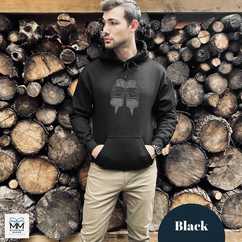 Op de afbeelding: Zwart hoodie met een afbeelding van een paar schaatsen. De hoodie wordt gedragen door een persoon die voor een muur van gestapelde boomstammen staat.