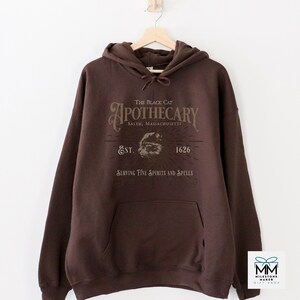 May include: A dark brown hoodie with a front pocket and a drawstring. The front displays the text "The Black Cat Apothecary Salem, Massachusetts Est. 1626 Serving Fine Spirits and Spells" in a vintage style.