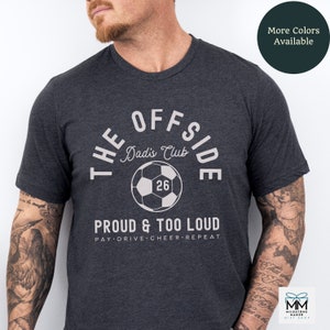 May include: A dark heather grey t-shirt with a white graphic design that says "The Offside Dad's Club" with a soccer ball and the number 26. The text below the graphic says "Proud & Too Loud" and "Pay Drive Cheer Repeat".