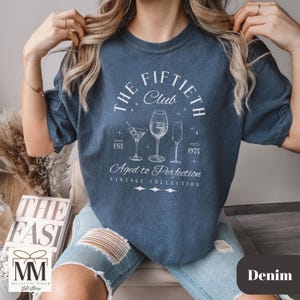 May include: Denim blue t-shirt with the text "The Fiftieth Club" and the words "Aged to Perfection". The shirt features illustrations of cocktail glasses and the year 1975. The shirt is part of a vintage collection.