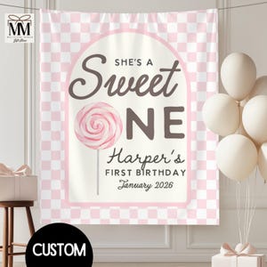 May include: A pink and white checkered backdrop with the text "SHE'S A Sweet ONE" and "Harper's FIRST BIRTHDAY January 2026". A large pink lollipop graphic is in the center. White balloons are on the right.
