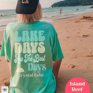 Może przedstawiać: Turkusowa koszulka z napisem "LAKE DAYS Are The Best Days" w stylu retro, ze słońcem i napisem "Crystal Lake" poniżej. Osoba ma na sobie czarną czapkę z napisem "LA" i siedzi na piaszczystej plaży.