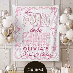 Pode incluir: Uma faixa branca com texto e detalhes em rosa para uma festa de primeiro aniversário. A faixa diz "it's FUN to be ONE" com o nome "Olivia's First Birthday" abaixo. Balões em prata e branco circundam a faixa.