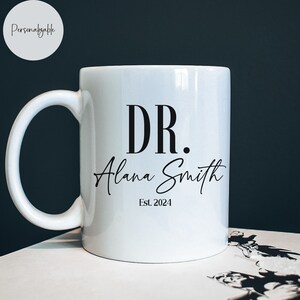 May include: White ceramic coffee mug with black text that reads "DR. Alana Smith Est. 2024".