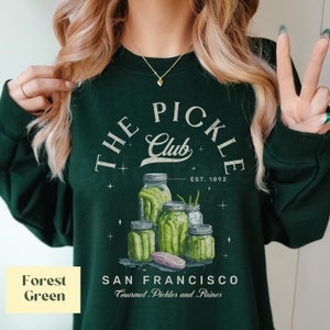 Puede incluir: Sudadera verde oscuro con un dise&ntilde;o gr&aacute;fico de tarros de pepinillos y el texto "THE PICKLE Club, EST. 1892, SAN FRANCISCO, Gourmet Pickles and Brines." El dise&ntilde;o presenta tarros de pepinillos de varios tama&ntilde;os.