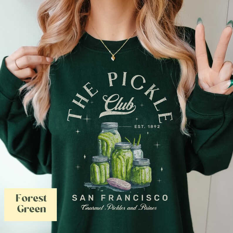 Puede incluir: Sudadera verde oscuro con un dise&ntilde;o gr&aacute;fico de tarros de pepinillos y el texto "THE PICKLE Club, EST. 1892, SAN FRANCISCO, Gourmet Pickles and Brines." El dise&ntilde;o presenta tarros de pepinillos de varios tama&ntilde;os.