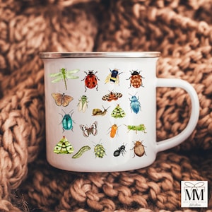 May include: White enamel mug featuring a collection of colorful insect illustrations, including a dragonfly, ladybug, and butterfly. The mug has a silver rim and a white handle. The background is a brown, textured fabric.
