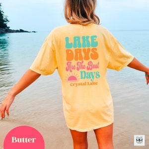 Może przedstawiać: Ż&oacute;łta koszulka z napisem "LAKE DAYS Are The Best Days" w stylu retro. Koszulka posiada r&oacute;wnież grafikę zachodu słońca oraz napis "Crystal Lake". Koszulka jest w kolorze maślanym ż&oacute;łtym.
