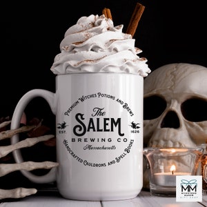 Puede incluir: Taza de cer&aacute;mica blanca con un dise&ntilde;o gr&aacute;fico en blanco y negro que presenta un caldero de bruja y el texto "The Salem Brewing Co. Massachusetts" con una bruja volando en una escoba a cada lado del texto. La taza tiene un asa negra y est&aacute; llena de crema batida y un palo de canela.