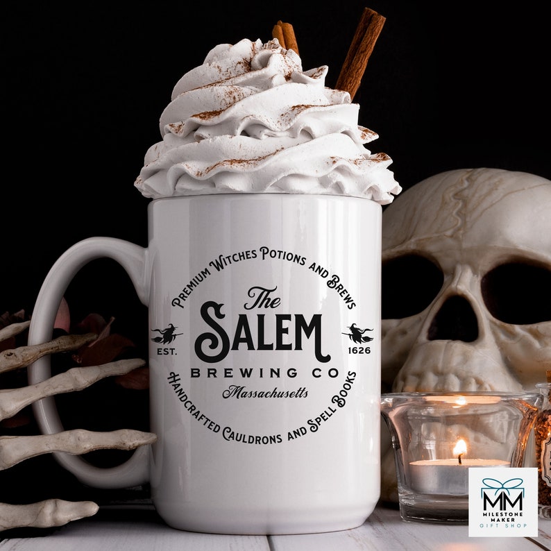Puede incluir: Taza de cer&aacute;mica blanca con un dise&ntilde;o gr&aacute;fico en blanco y negro que presenta un caldero de bruja y el texto "The Salem Brewing Co. Massachusetts" con una bruja volando en una escoba a cada lado del texto. La taza tiene un asa negra y est&aacute; llena de crema batida y un palo de canela.