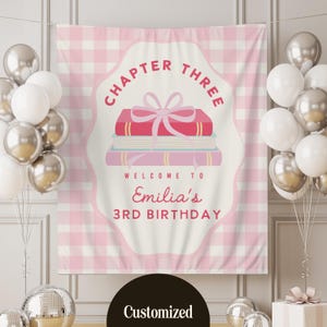 May include: A pink and white checkered backdrop with a banner that reads "Chapter Three" and "Welcome to Emilia's 3rd Birthday." The banner features a stack of books with a pink bow. Silver and white balloons are on either side.