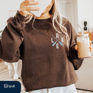 K&ouml;nnte beinhalten: Ein braunes Sweatshirt mit einer wei&szlig;en Grafik von gekreuzten Paddeln und dem Wort "LAKE" auf der Vorderseite.