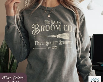 Puede incluir: Una sudadera gris con un diseño gráfico de estilo vintage que dice "The Salem Broom Co. Finest Quality Brooms Est. 1626". El gráfico presenta una escoba estilizada con un mango y cerdas.