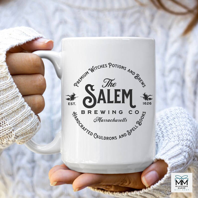 Puede incluir: Taza de cer&aacute;mica blanca con un dise&ntilde;o gr&aacute;fico en blanco y negro. El gr&aacute;fico presenta un caldero de bruja estilizado y el texto "The Salem Brewing Co. Massachusetts. Premium Witches Potions and Brews. Est. 1626. Handcrafted Cauldrons and Spell Books."