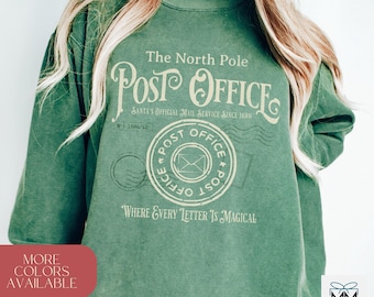以下が含まれることがあります： 「The North Pole Post Office Santa's Official Mail Service Since 1686」と、切手デザインと「Where Every Letter Is Magical」というテキストが書かれたヴィンテージスタイルのグラフィックが施された緑色のスウェットシャツ。
