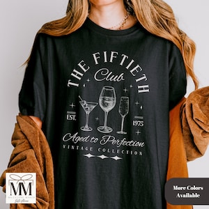 May include: Black t-shirt with a white graphic that reads "The Fiftieth Club" with wine glasses and the text "Aged to Perfection". The shirt also has the text "EST. 1975" and "Vintage Collection".