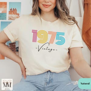 以下が含まれることがあります： 薄いベージュのTシャツに、虹色の1975のグラフィックと、その下に「ヴィンテージ」という文字が書かれています。モデルは青いジーンズを履いています。