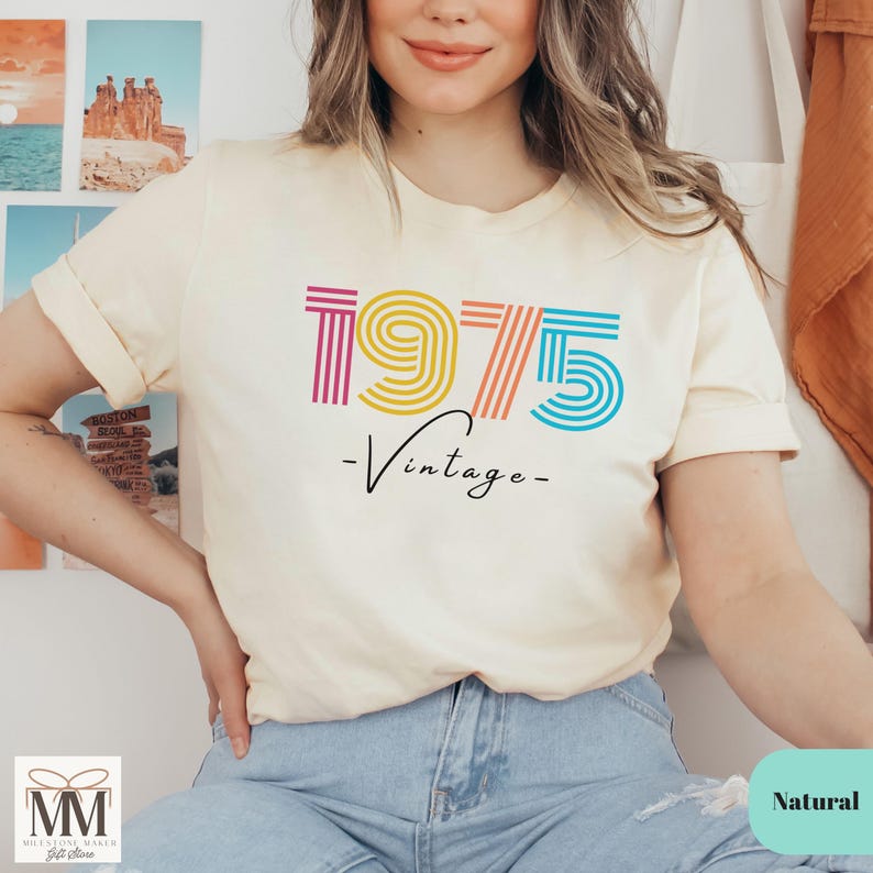 以下が含まれることがあります： 薄いベージュのTシャツに、虹色の1975のグラフィックと、その下に「ヴィンテージ」という文字が書かれています。モデルは青いジーンズを履いています。
