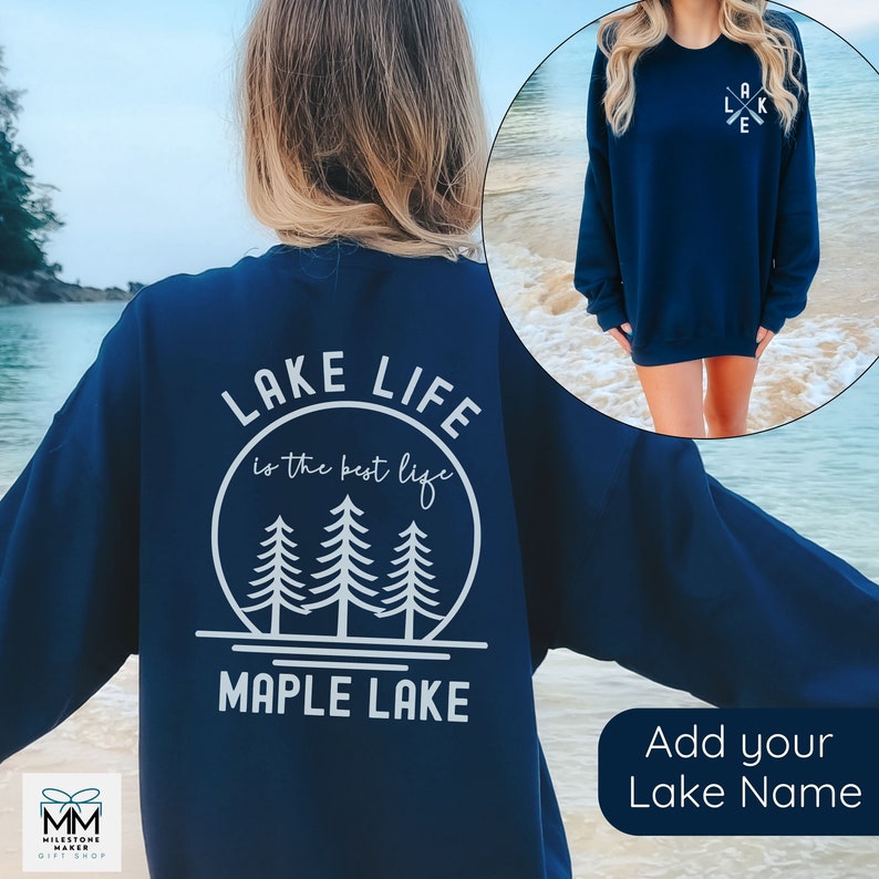 K&ouml;nnte beinhalten: Navyblauer Sweatshirt mit einem wei&szlig;en Grafikdesign auf der R&uuml;ckseite, das "Lake Life is the best life" mit drei Kiefern in einem Kreis und "Maple Lake" unter dem Kreis sagt. Die Vorderseite des Sweatshirts hat ein wei&szlig;es Grafikdesign aus zwei gekreuzten Rudern mit dem Wort "Lake" in der Mitte.