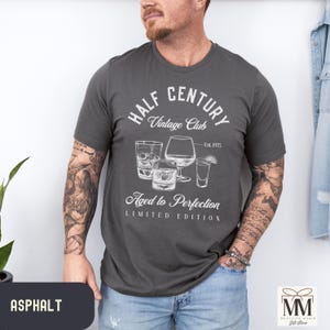 May include: Dark grey t-shirt featuring a white graphic with the text "HALF CENTURY Vintage Club" and "Aged to Perfection, Limited Edition." The design includes illustrations of cocktail glasses. A casual wear item, the shirt is a dark grey colour.