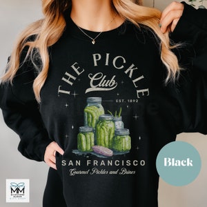 Puede incluir: Sudadera negra con un gr&aacute;fico de tarros de pepinillos y el texto "THE PICKLE Club EST. 1892 SAN FRANCISCO Gourmet Pickles and Brines." El dise&ntilde;o incluye pepinillos verdes y un pepinillo morado.