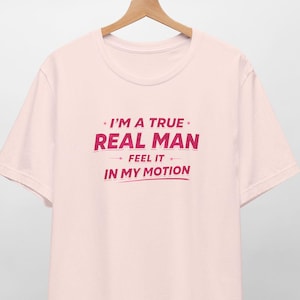 May include: A pale pink t-shirt featuring the burgundy text: "I'M A TRUE REAL MAN - FEEL IT - IN MY MOTION". The tee is suspended on a wooden hanger against a white backdrop.