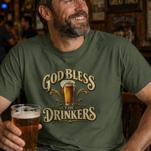 May include: An olive green short-sleeve t-shirt featuring a graphic print of a beer glass with foam. The text reads "GOD BLESS THE DRINKERS" in a decorative, arched font. The design includes ornate, swirling flourishes surrounding the beer glass.