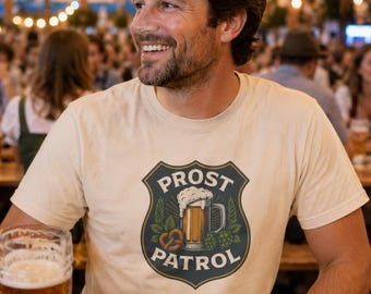 May include: A cream-colored short-sleeve t-shirt featuring a dark shield-shaped graphic on the chest. The graphic displays a beer mug with foam, a pretzel, and hop cones, with the text PROST PATROL arched around the central illustration.