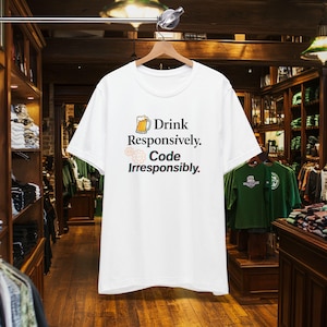 Pode incluir: Camiseta branca com o texto "Drink Responsibly. Code Irresponsibly." em preto e amarelo. O design inclui um caneco de cerveja e um ícone de engrenagem. A camisa está pendurada em um cabide de madeira em uma loja de roupas.
