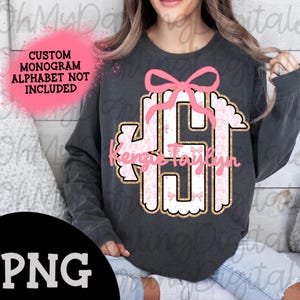 May include: A dark gray sweatshirt with a pink and gold monogram design. The monogram is surrounded by a white scalloped border and features a pink bow. The text "Kenzie Taylyn" is written below the monogram.