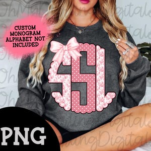May include: A gray sweatshirt with a pink monogram design featuring a bow. The monogram is surrounded by a white floral pattern and pink polka dots. The text "CUSTOM MONOGRAM ALPHABET NOT INCLUDED" is visible in the top left corner.