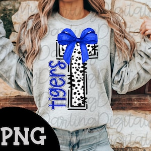 May include: A light gray sweatshirt featuring a cross design with a blue bow at the top. The cross has a black and white spotted pattern. The word "Tigers" is written vertically in blue on the left side of the cross.
