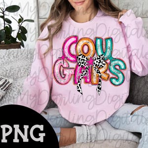 May include: A light pink sweatshirt with the word "COUGARS" in a colorful, distressed font. The design includes a bow with a black and white cow print and star accents. The sweatshirt is a casual style.