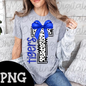 May include: A gray t-shirt featuring a cross design with a blue bow and a black and white dalmatian print. The word "Tigers" is written vertically in blue. The image has a PNG file format.