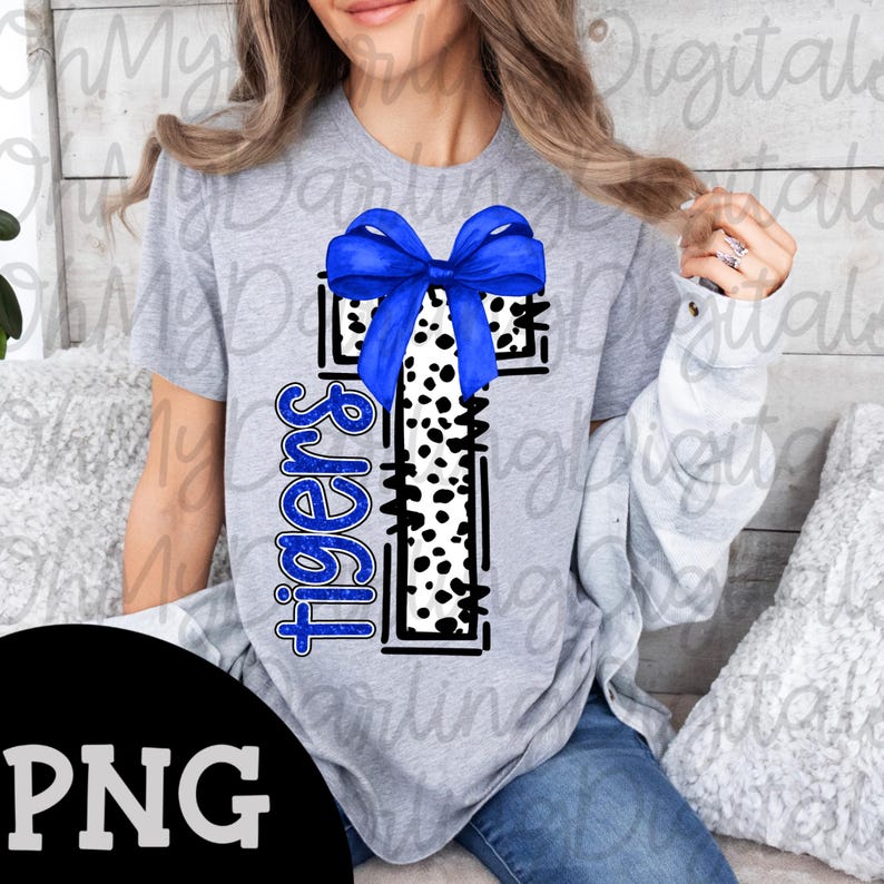 May include: A gray t-shirt featuring a cross design with a blue bow and a black and white dalmatian print. The word "Tigers" is written vertically in blue. The image has a PNG file format.