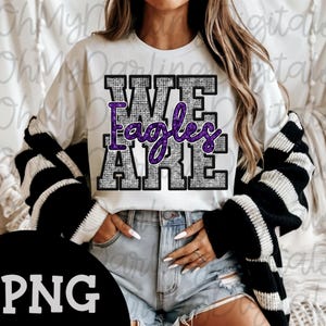 May include: White t-shirt with the text "WE ARE Eagles" in a textured, gray and purple design. The shirt is paired with denim shorts and a black and white striped cardigan.