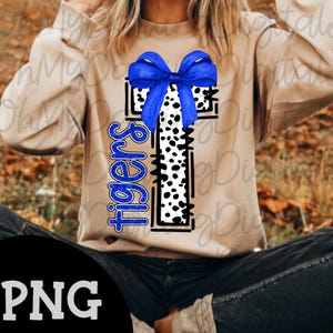 May include: A tan sweatshirt featuring a large, stylized letter "T" with a black and white dalmatian print. A blue bow sits atop the "T", and the word "Tigers" is written vertically in blue. The image has a PNG watermark.