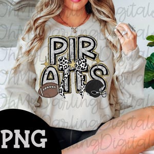 May include: A beige sweatshirt featuring the word "PIRATES" in a distressed black and white font, with gold edging. The design includes a football, a helmet, and a bow with a black and white spotted pattern. Two gold stars complete the design.