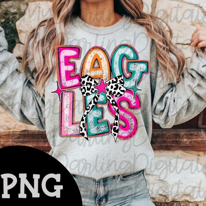 May include: A gray sweatshirt with the word "EAGLES" in colorful, bold letters. The letters are outlined in pink, teal, and orange, with a black and white bow in the center. Pink star accents are on either side.