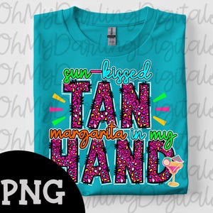 以下が含まれることがあります： 「sun-kissed TAN margarita in my HAND」の文字が入ったターコイズ色のTシャツ。 「TAN」の文字はピンクのヒョウ柄です。 カクテルのグラフィックが右下にあります。 シャツは折りたたまれています。