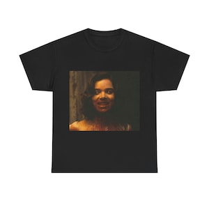 Puede incluir: Una camiseta negra con una imagen gráfica de una mujer con sangre en la cara y el pecho. La mujer sonríe.