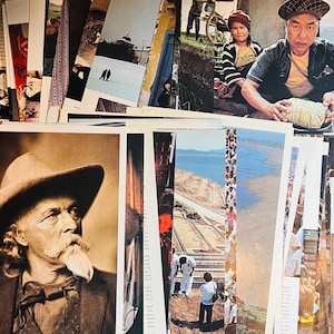 May include: A collection of vintage postcards featuring a variety of subjects, including a man in a hat, a landscape with a river, and a group of people. The postcards are in various colors and sizes, and some have handwritten notes on the back.