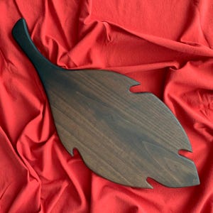 Puede incluir: Una tabla de servir de madera marrón oscuro con forma de hoja y un mango largo. La tabla tiene una superficie lisa y pulida y es perfecta para servir queso, embutidos u otros aperitivos.