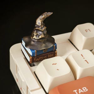 Pode incluir: Um chapéu de seleção em miniatura da série Harry Potter, feito de resina pintada de marrom e dourado, está em cima de uma pilha de três livros em miniatura. Os livros são azuis, vermelhos e marrons com letras douradas que dizem "Sorting Hat". O conjunto em miniatura está em cima de um teclado branco.