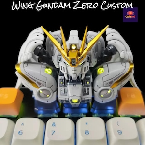 May include: A white, gold, and blue Wing Gundam Zero Custom model, with the text "WING GUNDAM ZERO CUSTOM" above it. The model is placed on a keyboard with white and orange keys. The model has a detailed design with glowing accents.