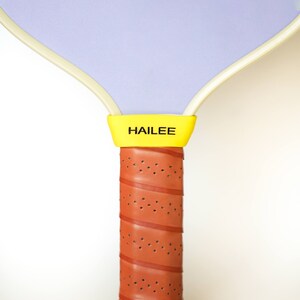 Puede incluir: Una raqueta de paddleball morada y blanca con un mango amarillo y la palabra "HAILEE" impresa en el mango.