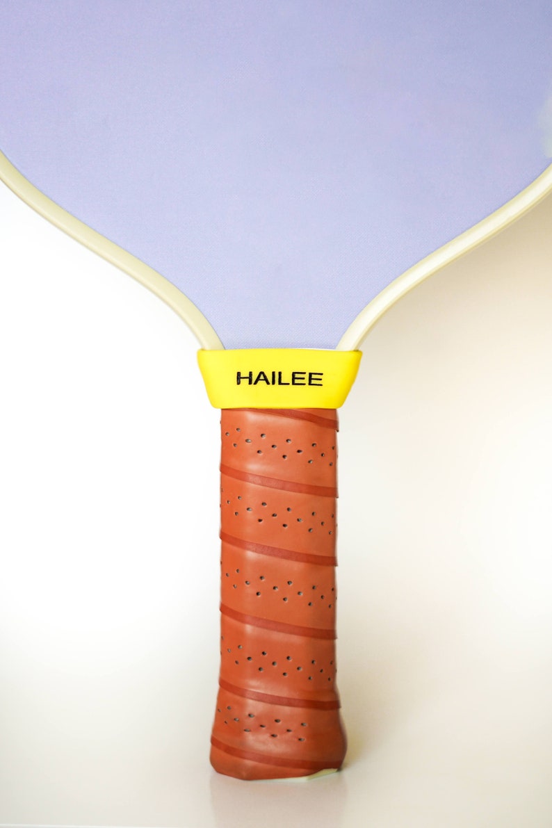 Puede incluir: Una raqueta de paddleball morada y blanca con un mango amarillo y la palabra "HAILEE" impresa en el mango.