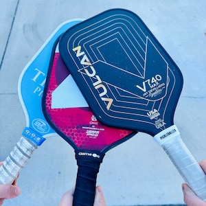 Op de afbeelding: Close-up van drie pickleball rackets. E&eacute;n is lichtblauw, een andere donkerrood en de derde zwart met gouden details. De rackets worden door handen vastgehouden, waardoor hun grepen zichtbaar zijn. Voor de sport pickleball.
