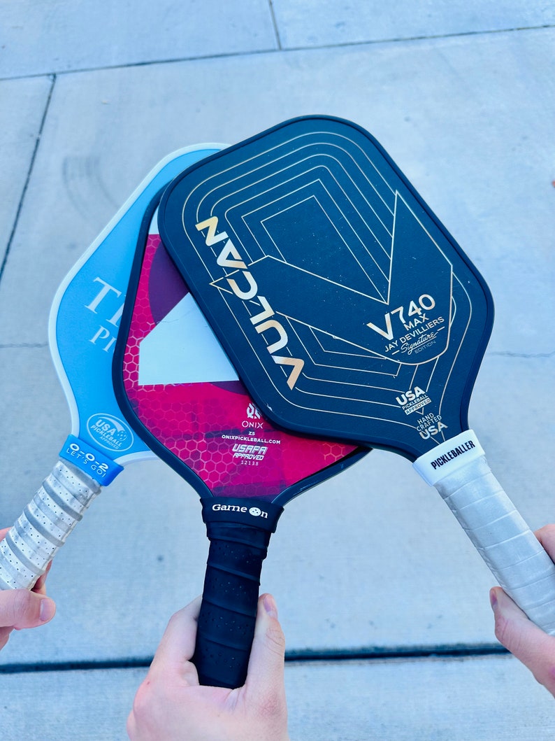 Op de afbeelding: Close-up van drie pickleball rackets. E&eacute;n is lichtblauw, een andere donkerrood en de derde zwart met gouden details. De rackets worden door handen vastgehouden, waardoor hun grepen zichtbaar zijn. Voor de sport pickleball.