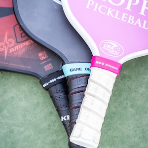 Puede incluir: Tres palas de pickleball con diferentes dise&ntilde;os y colores. La pala superior es rosa con texto blanco que dice "Trophy Pickleball". La pala del medio es negra con texto blanco que dice "Quality". La pala inferior es roja y negra con texto blanco que dice "Amped Midweight".